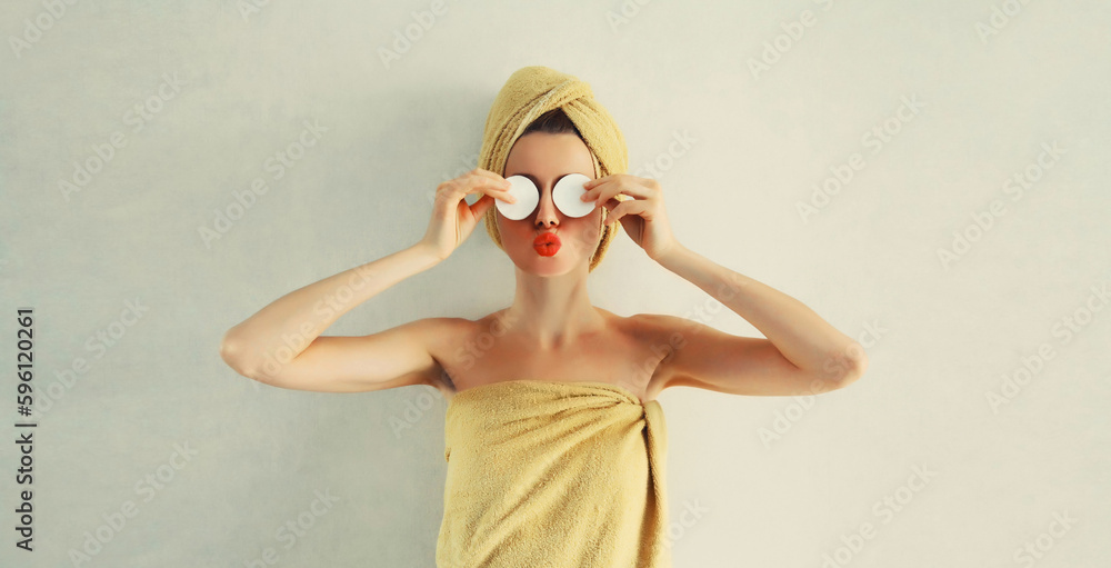 Portrait of happy young caucasian woman with cotton pads applying face cream and drying wet hair in wrapped bath towel