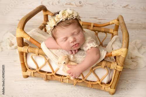 A cute little newborn baby in a white suit and a headband with flowers on his head sleeps sweetly. Wicker rattan bed with white linens. Professional macro photo against a light wood background.