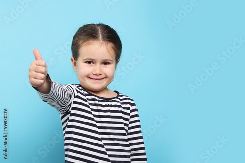 Happy girl showing thumbs up on light blue background. Space for text