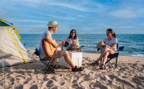 Group of happy Asian friends playing guitar and singing with clapping together having fun and enjoying while camping picnic on the beach in summer vacation.