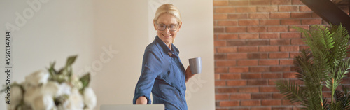 Cheerful blonde mature woman using laptop computer while getting ready for working day, standing in modern kitchen room photo