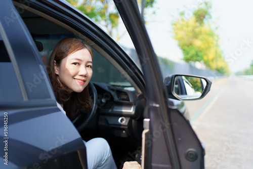 Happy cheerful Asian woman getting in or getting out of the car from driver position, female driver smiles and looks at camera.