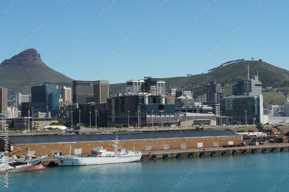 Cityscape of Cape Town with modern buildings and on background is Lion's Head and Signal Hill under blue sky observed from port. In foreground are fishing vessels.