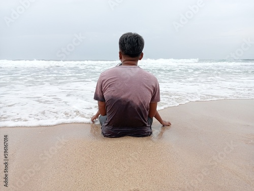 A man sits on the edge of a white sandy beach and looks out to sea.