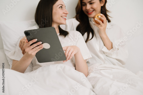 Attractive lesbian couple use mobile phone, tablet watch movie on bed. Beautiful sibling in pajamas lying down with blanket in bedroom together, in morning.