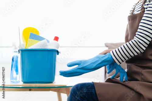 Woman in apron puts on rubber gloves and ready to housework sitting in living room interior. Housewife with cleaning equipment has fun and many household chores, professional cleaning service.
