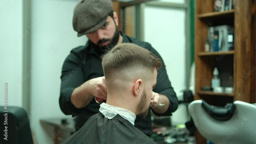 Trendy barber cuts bearded man's hair with a clipper in barbershop. Men's hairstyling and hair cutting in salon. Grooming the hair with trimmer. Hairdresser doing haircut in retro hair salon. Trendy barber cuts bearded man's hair with a clipper in barbershop. Men's hairstyling and hair cutting in salon. Grooming the hair with trimmer. Hairdresser doing haircut in retro hair salon.