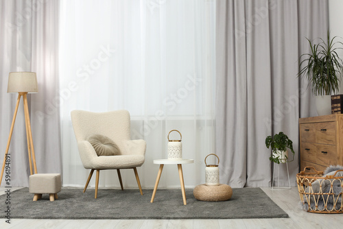 Comfortable armchair, white table and stylish lanterns near elegant curtains in light room. Interior design