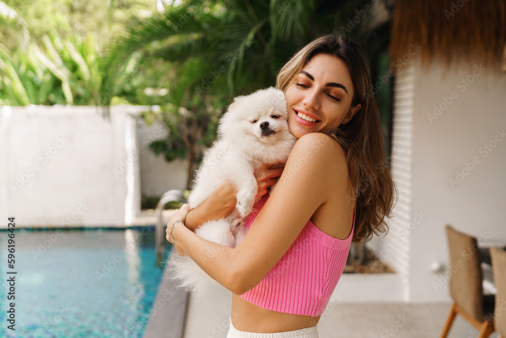 Pretty brunette woman with wavy hairs hugging with cute playful pomeranian puppy in her own stylish villa with pool. Pretty brunette woman with wavy hairs hugging with cute playful pomeranian puppy in her own stylish villa with pool.
