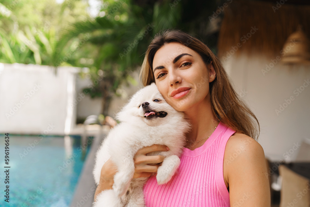 Adorable toy pomeranian dog in arms of its loving owner. Small adorable doggy with funny fur with pretty woman. Tropical moder villa on background. Adorable toy pomeranian dog in arms of its loving owner. Small adorable doggy with funny fur with pretty woman. Tropical moder villa on background.