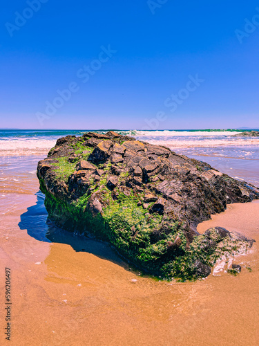 Vertical Shot of rock formation with vibrant green moss growing on the sides splashed in foaming white ocean waves on a warm sandy beach with turquoise blue green ocean shimmering in the background