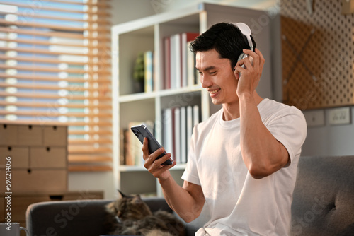 Happy Asian man enjoy favorite songs in headphones and using smartphone on couch. Technology and lifestyle concept