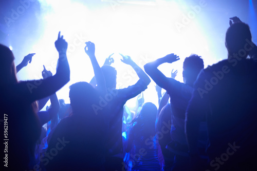 Blue, neon lights and people dancing at music festival from back, lighting and energy at live concert event. Dance, fun and group of excited fans in arena at rock band performance or crowd at party.