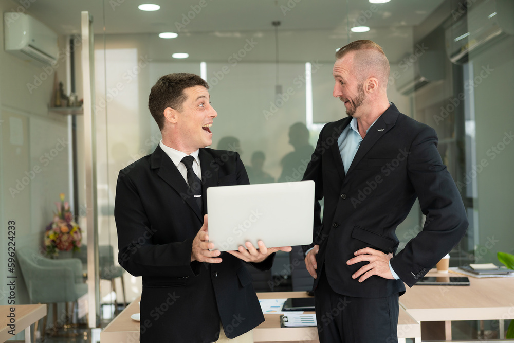 Two young businessmen looking at laptop smiling and expressing joy that the business succeeded according to their plans colleague talking to each other describing good partnership
