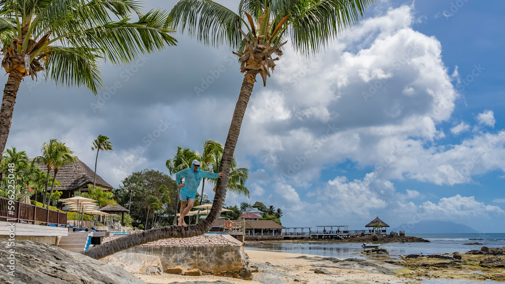 Obraz premium A palm tree leaned over the beach. A man in sunglasses and a cap is standing on a curved trunk. In the distance - the buildings of the hotel, the ocean at low tide. Clouds in the sky. Seychelles. Mahe