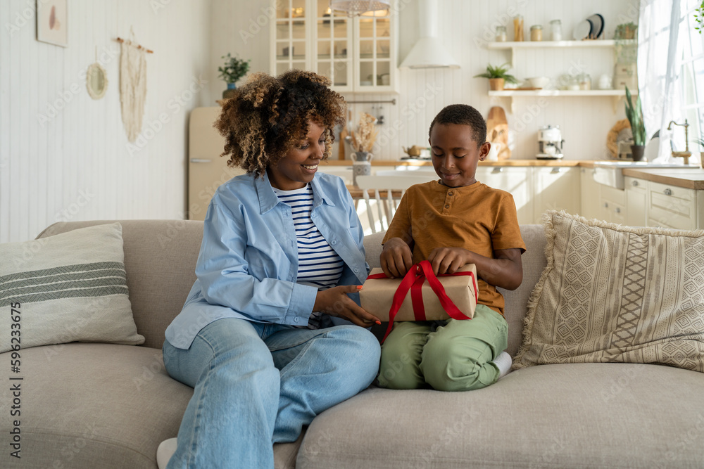 Happy African American child boy opening wrapped birthday gift while sitting on sofa with mother at home. Excited kid untying red ribbon on holiday present, celebrating special family occasions Happy African American child boy opening wrapped birthday gift while sitting on sofa with mother at home. Excited kid untying red ribbon on holiday present, celebrating special family occasions