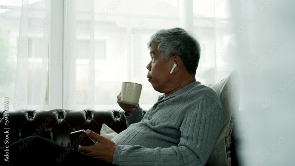 Active senior man relaxing at home working, wearing bluetooth earbud enjoy listening to music from a mobile phone sitting comfortably on a sofa. Active Senior concept. Active senior man relaxing at home working, wearing bluetooth earbud enjoy listening to music from a mobile phone sitting comfortably on a sofa. Active Senior concept.