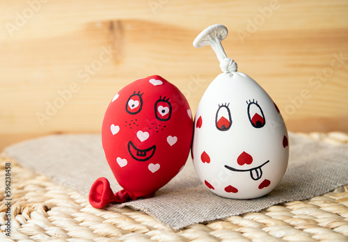 Two funny homemade sensory stress balls, made of balloons and filled with flour. Red and white balls with hearts and funny drawn faces, wood background.