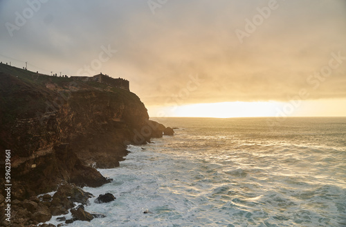 View of the sunset and the old lighthouse on a rock with a fortress on the coast of the Atlantic Ocean in the city of Nazare, Portugal. High quality photo