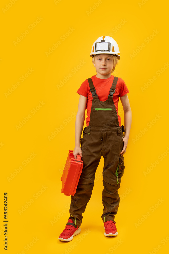 Boy imitates worker. Child in work overalls and helmet with suitcase of tools. Mock up, copy space. Vertical frame Boy imitates worker. Child in work overalls and helmet with suitcase of tools. Mock up, copy space. Vertical frame