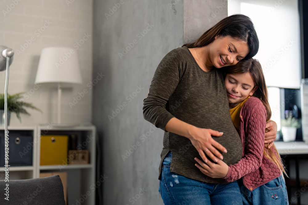 Cheerful moments of smiling pregnant single mother with young elementary age daughter at home. Cute young girl embracing her mother and tenderly touching her stomach. Cheerful moments of smiling pregnant single mother with young elementary age daughter at home. Cute young girl embracing her mother and tenderly touching her stomach.