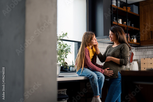 Beautiful smiling pregnant woman and her daughter enjoying while embracing in kitchen at home.