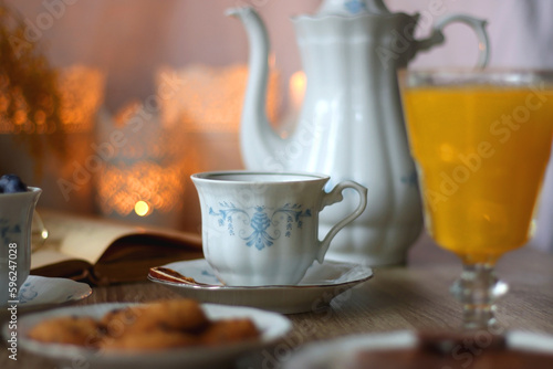 Cup of tea or coffee, plate of cookies, cup of blueberries, plate of chocolate, glass of juice, book, reading glasses, teapot, flowers and lit candles on the table. Brunch or afternoon tea concept.