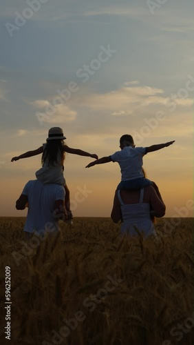 Vertical Screen: Slow motion mom and dad carrying kids on shoulders while walking in wheat field at sunset, boy and girl spreading arms as if flying, blue sky on background. Happy family together