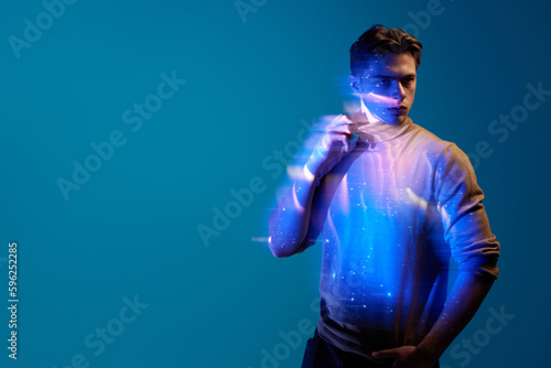 Handsome, stylish, young man with serious expression posing with digital kosmos reflection on body and clothes against blue background in neon. Modern photography, art, cyberpunk, creativity concept