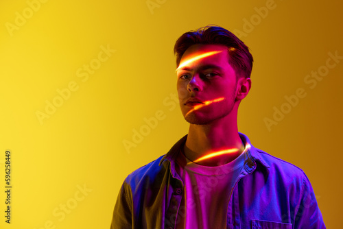 Handsome young man in casual stylish clothes with digital neon filter reflection on face standing against gradient yellow background. Concept of modern photography, art, cyberpunk, techno, creativity
