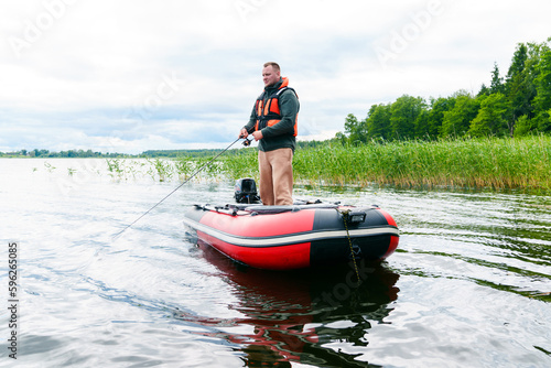 A man is fishing standing up from an inflatable boat. The lake is in clear water. Hobbies and recreation
