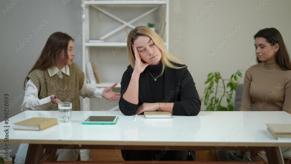 Sad tired therapist looking at camera as anxious angry teen girl and young woman arguing at background. Exhausted Caucasian woman posing indoors on appointment with irritated patients Sad tired therapist looking at camera as anxious angry teen girl and young woman arguing at background. Exhausted Caucasian woman posing indoors on appointment with irritated patients