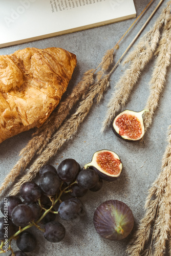 Autumn attributes. Croissant, dark grapes, book and figs on the grey table. Gold October