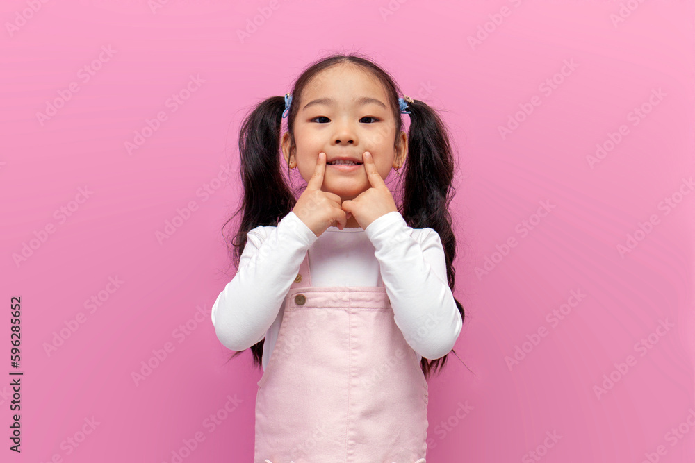 little Asian girl in pink sundress with long hair points her fingers at smile and healthy teeth, Korean preschool child little Asian girl in pink sundress with long hair points her fingers at smile and healthy teeth, Korean preschool child