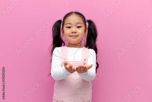 Obraz na płótnie little asian girl in pink sundress holds empty hands in front of her on pink iso
