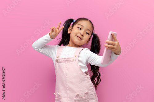 Obraz na plátně little asian girl in pink sundress takes selfie on smartphone and shows gesture