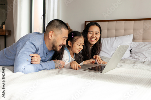 asian family with little daughter lie on bed at home and use laptop, korean dad mom and daughter look at computer screen