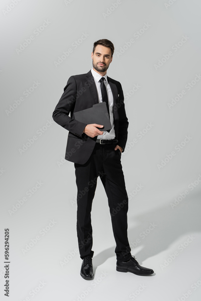 full length of successful and confident businessman in black stylish suit standing with folder on grey background. full length of successful and confident businessman in black stylish suit standing with folder on grey background.