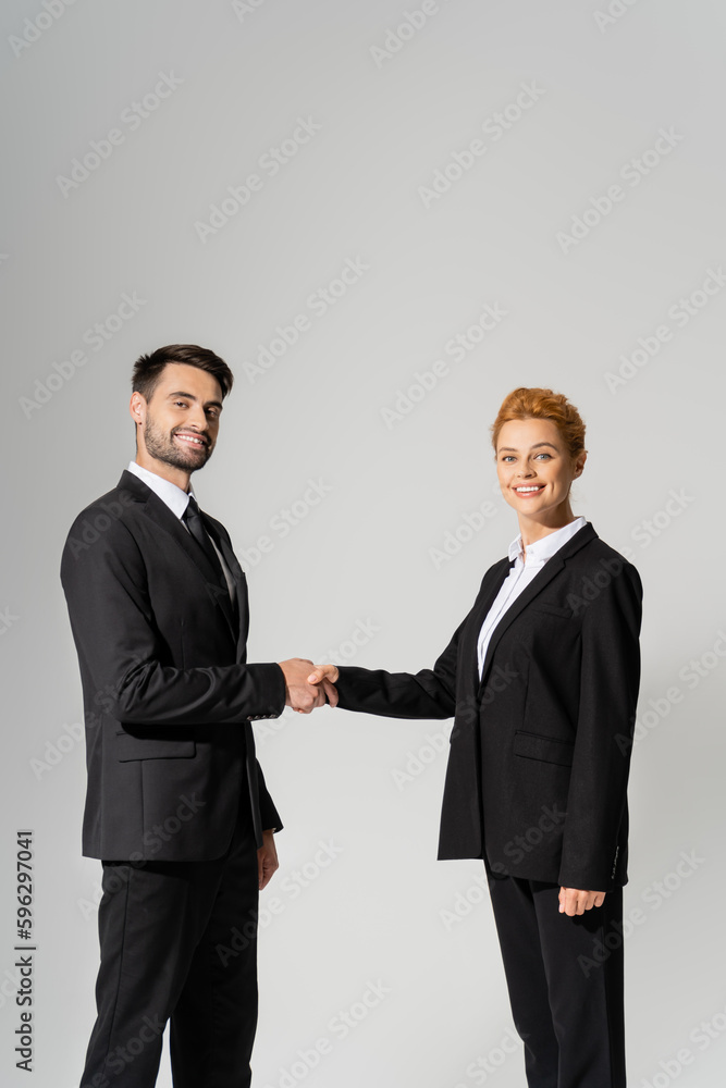 successful business partners in black blazers shaking hands and looking at camera isolated on grey.
