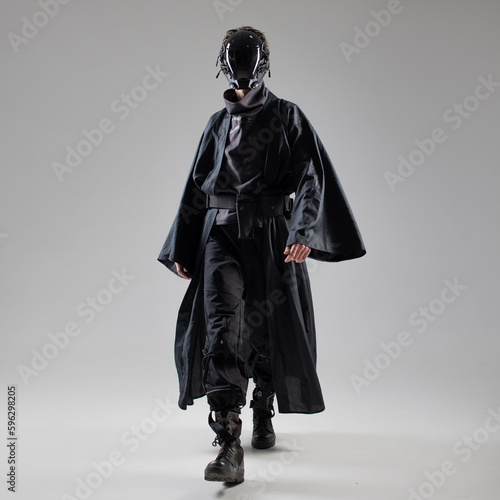 Urbantech outfit cyber style, a young man in stylish black clothes and a mirror mask on the whole face, futuristic style. Studio photo on a light background