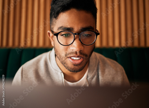 .. Closeup of geeky young indian man wearing glasses while reading something interesting and sitting inside. Man wearing glasses while reading online. Dedicated male student doing research in cafe. photo