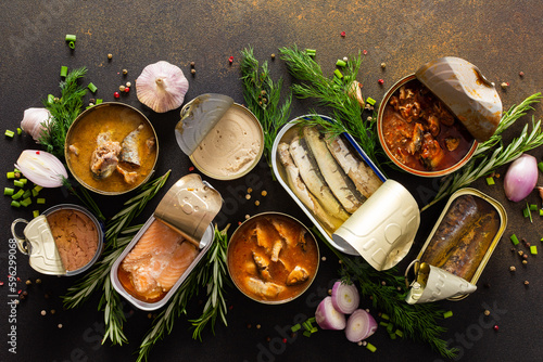 Different open tin cans with canned fish among spices and herbs on a brown background, canned salmon and mackerel, sprat and sardine, tuna and herring and fish pate, top view photo