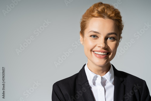 portrait of overjoyed businesswoman with red hair smiling at camera isolated on grey.