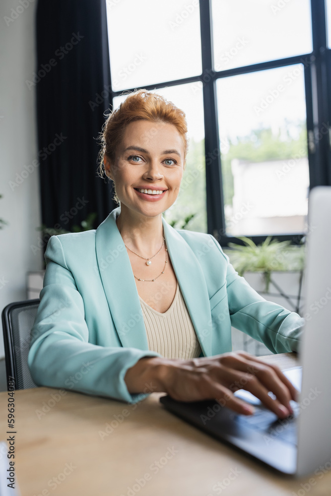 successful businesswoman in stylish blazer typing on laptop and smiling at camera in office. successful businesswoman in stylish blazer typing on laptop and smiling at camera in office.