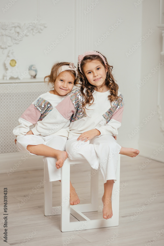 Two sisters in white sweatshirts are sitting on a chair and having fun and hugging. Two sisters in white sweatshirts are sitting on a chair and having fun and hugging.