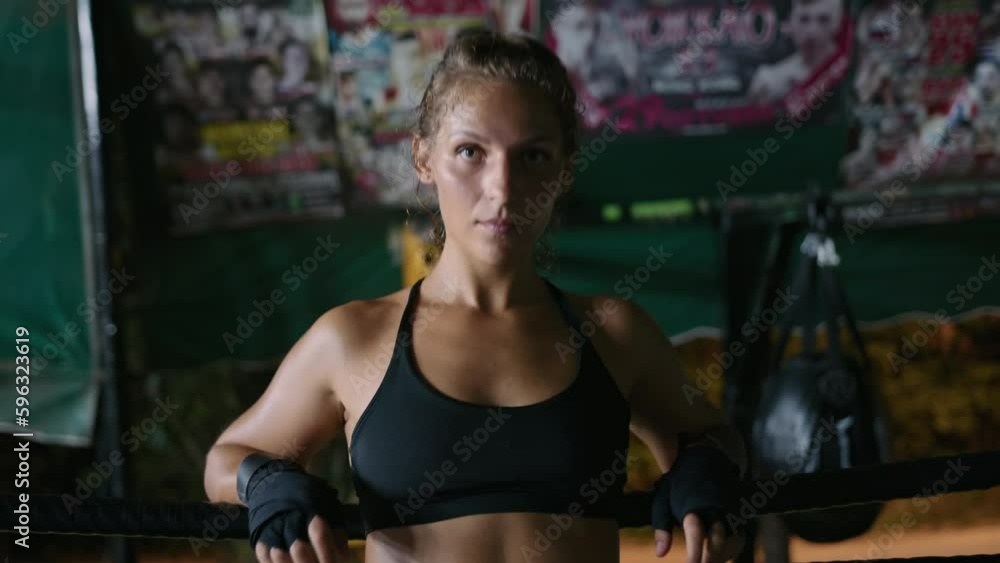 Portrait of woman fighter resting on ring ropes in gym and looking at camera. Female caucasian boxer with curly hair restores breathing between rounds at training session Portrait of woman fighter resting on ring ropes in gym and looking at camera. Female caucasian boxer with curly hair restores breathing between rounds at training session