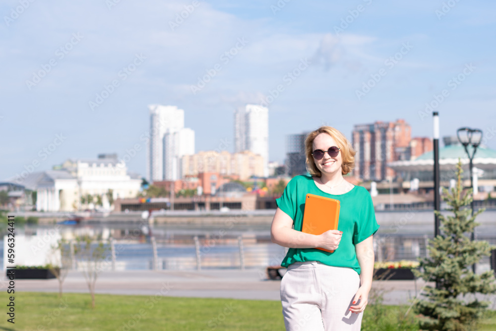 A smiling blogger girl in sunglasses holding a notebook or tablet in her hands on the embankment, on the street.