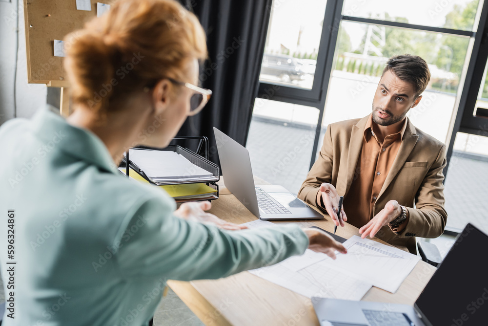 irritated business colleagues pointing at documents near laptops while quarreling in office. irritated business colleagues pointing at documents near laptops while quarreling in office.