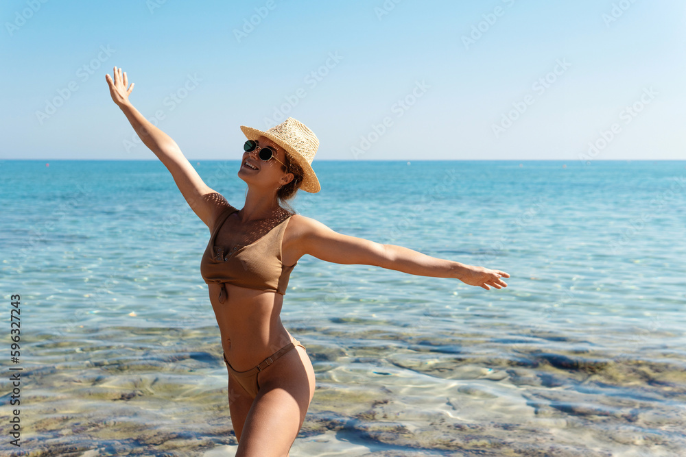 Carefree beautiful woman wearing brown bikini and straw hat during her beach vacation near blue sea