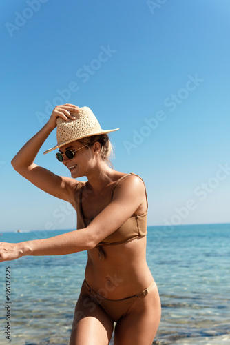Carefree beautiful woman wearing brown bikini and straw hat during her beach vacation near blue sea © blackday